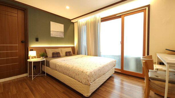 A bedroom with a bed, bedside table, lamp, and a view of the outside through a window. - OYO Rooftop Hostel (Seoul)