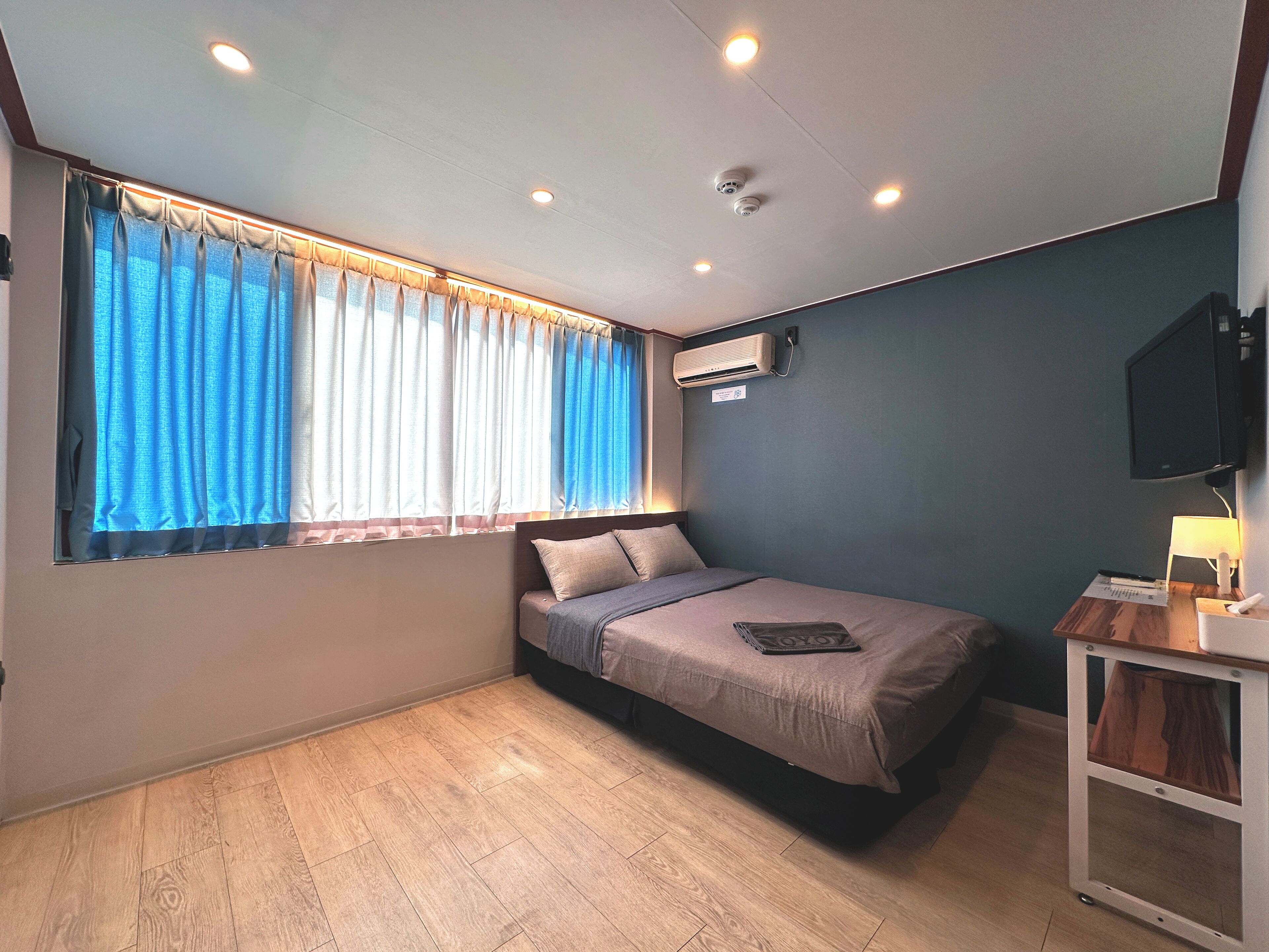 A compact bedroom with a bed, a window with curtains, an air conditioning unit, a TV mounted on the wall, and a bedside table with a lamp.