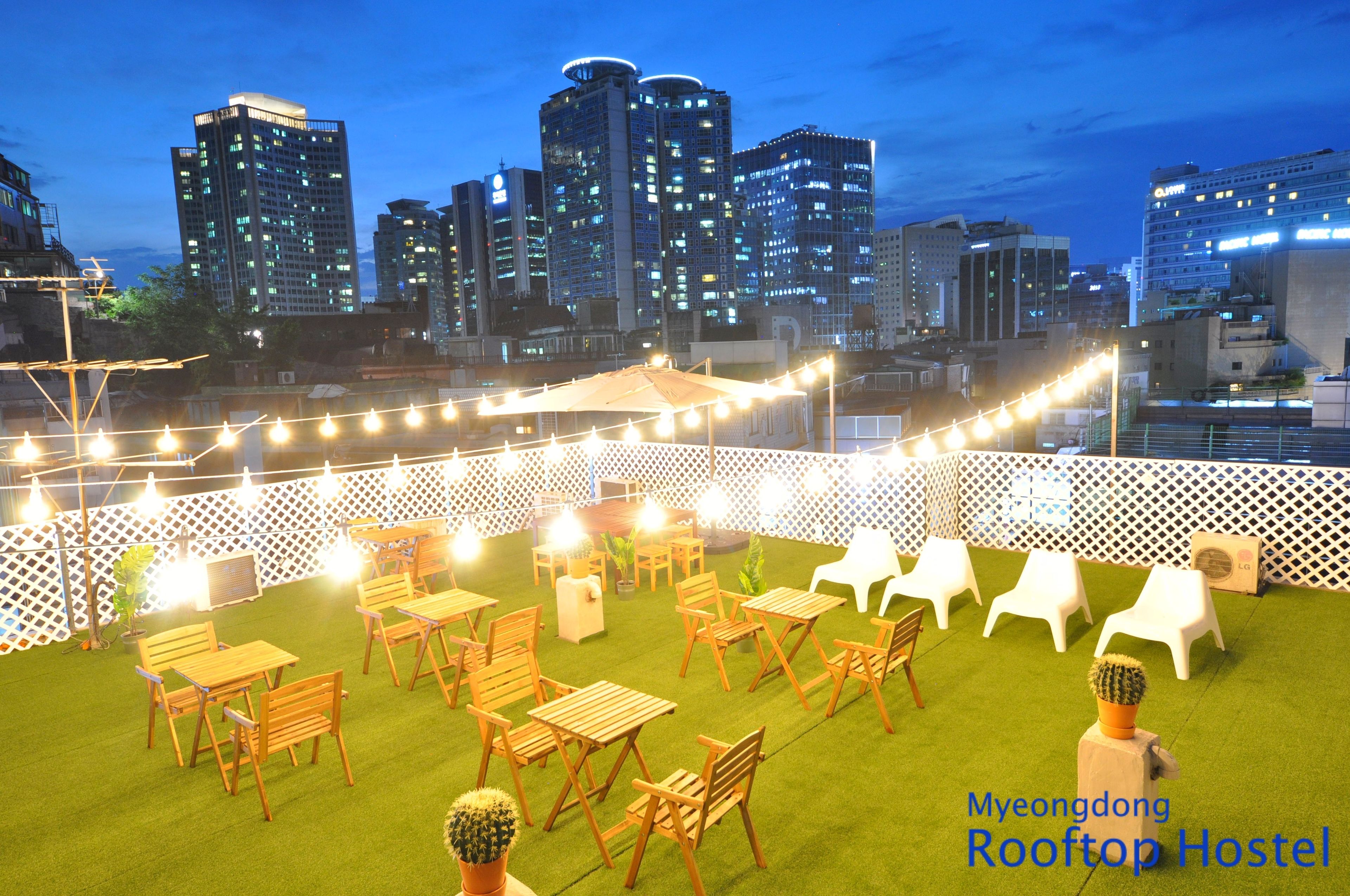 A rooftop hostel with wooden tables and chairs, string lights, and a view of the city skyline at dusk.