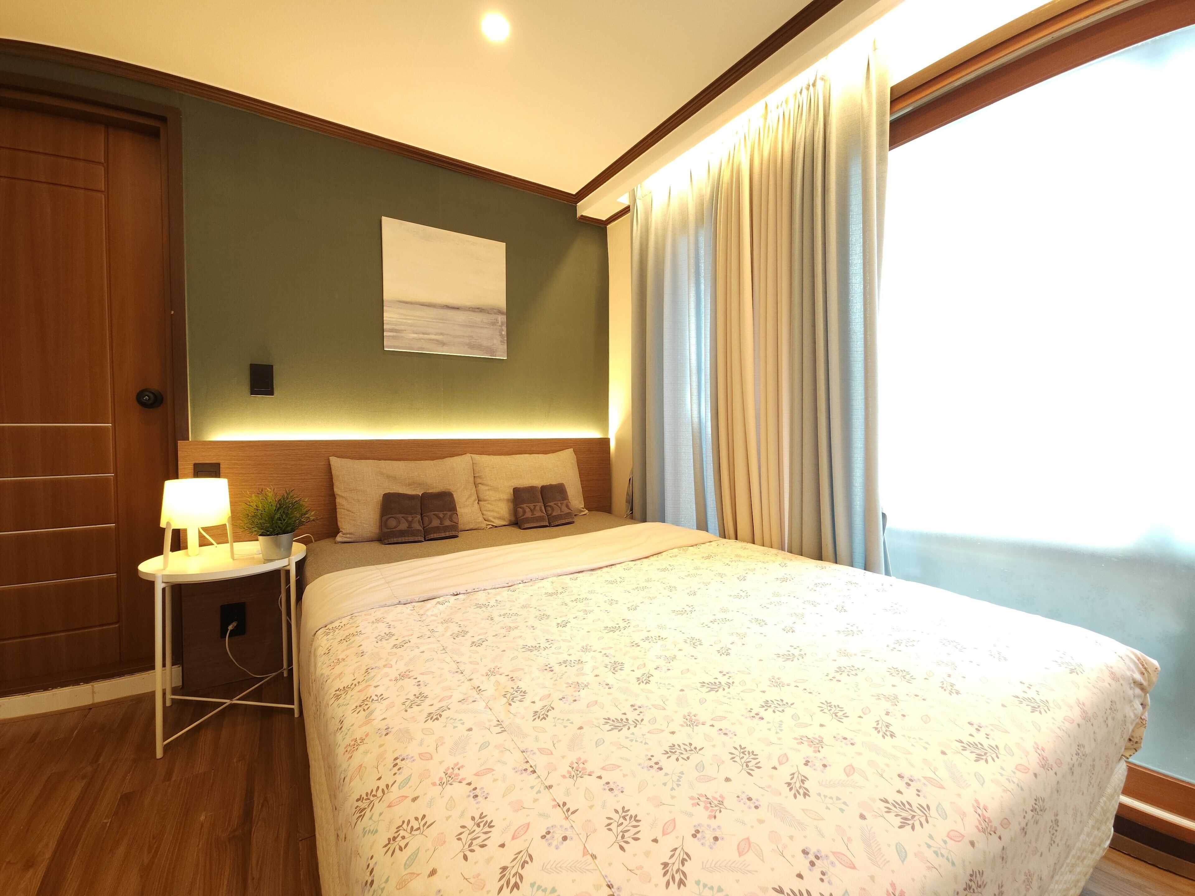 Une chambre d’hôtel avec un grand lit, une table de chevet avec une lampe, une plante en pot et une vue sur l’extérieur par une fenêtre avec des rideaux.