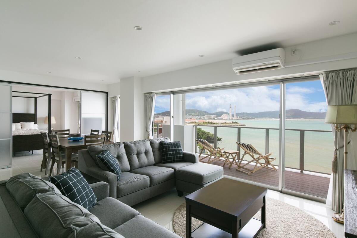 Ein modernes Wohnzimmer mit einem grauen Ecksofa, einem Esstisch und Stühlen. Ein großes Fenster bietet Blick auf das Meer und eine entfernte Skyline.