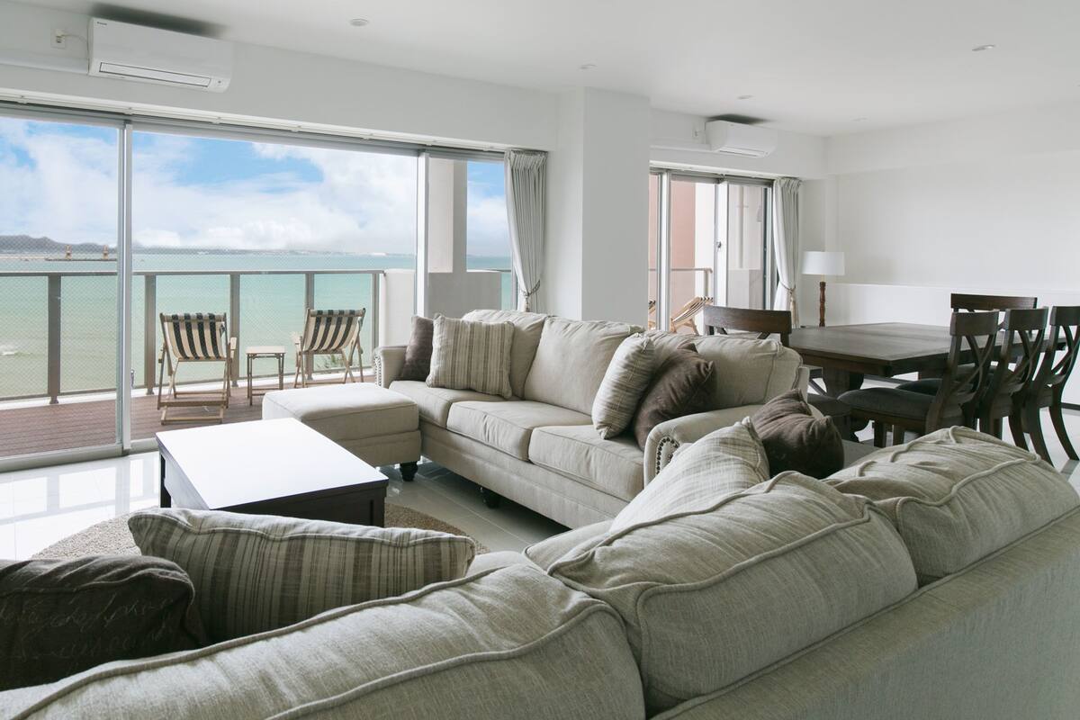 Ein geräumiges Wohnzimmer mit einer großen Couch, einem Couchtisch und einem Fernseher. Ein Balkon mit Blick aufs Meer.