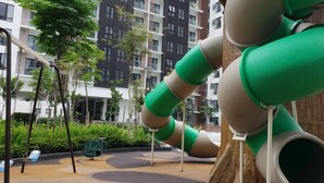 Un terrain de jeux avec un toboggan vert et beige, entouré d’arbres et avec un bâtiment visible au fond.
