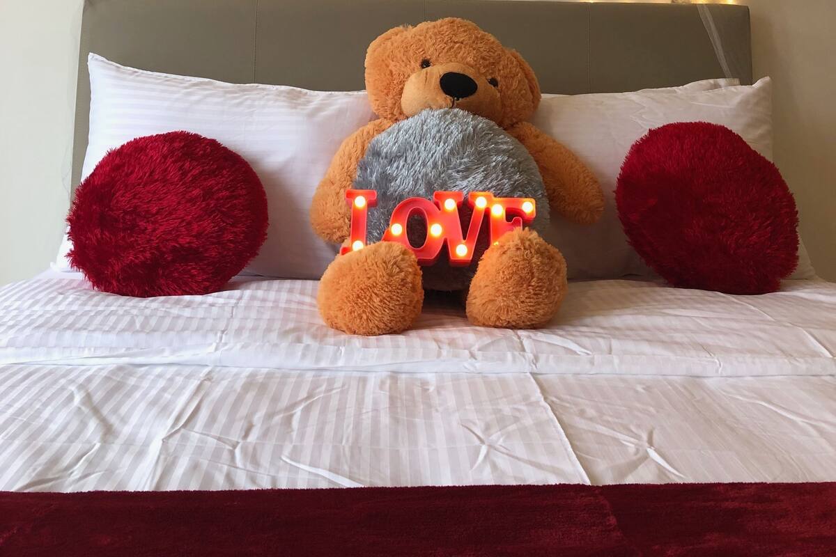 A bed with a teddy bear, red pillows, and a wall with 'LOVE' and 'IF YOU WOULD BE LOVABLE' text.