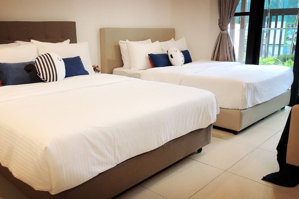 Two double beds with white linens and a variety of pillows, a brown headboard, and a view of greenery through a window.