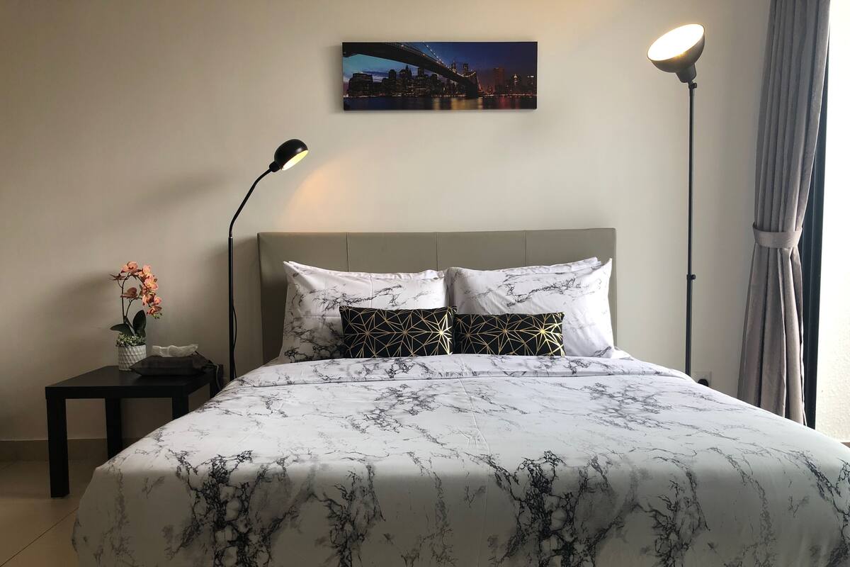 A neatly made bed with a marble-patterned bedspread, a cityscape painting above it, a floor lamp, and a small table with a potted orchid beside the bed.