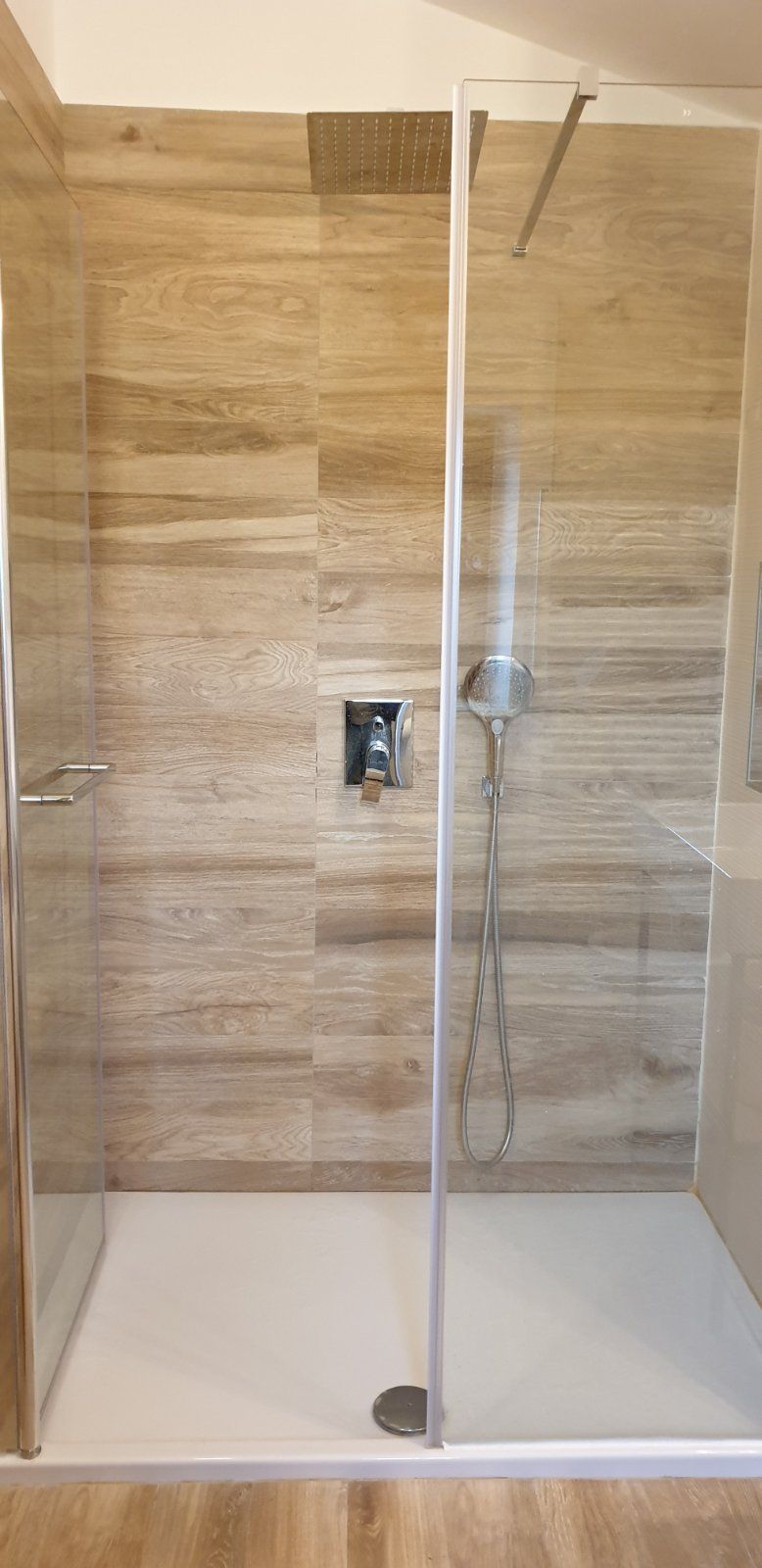 En dusch med glasväggar, en väggpanel i trä, ett duschmunstycke som kan hållas i handen och en kontrollpanel.
