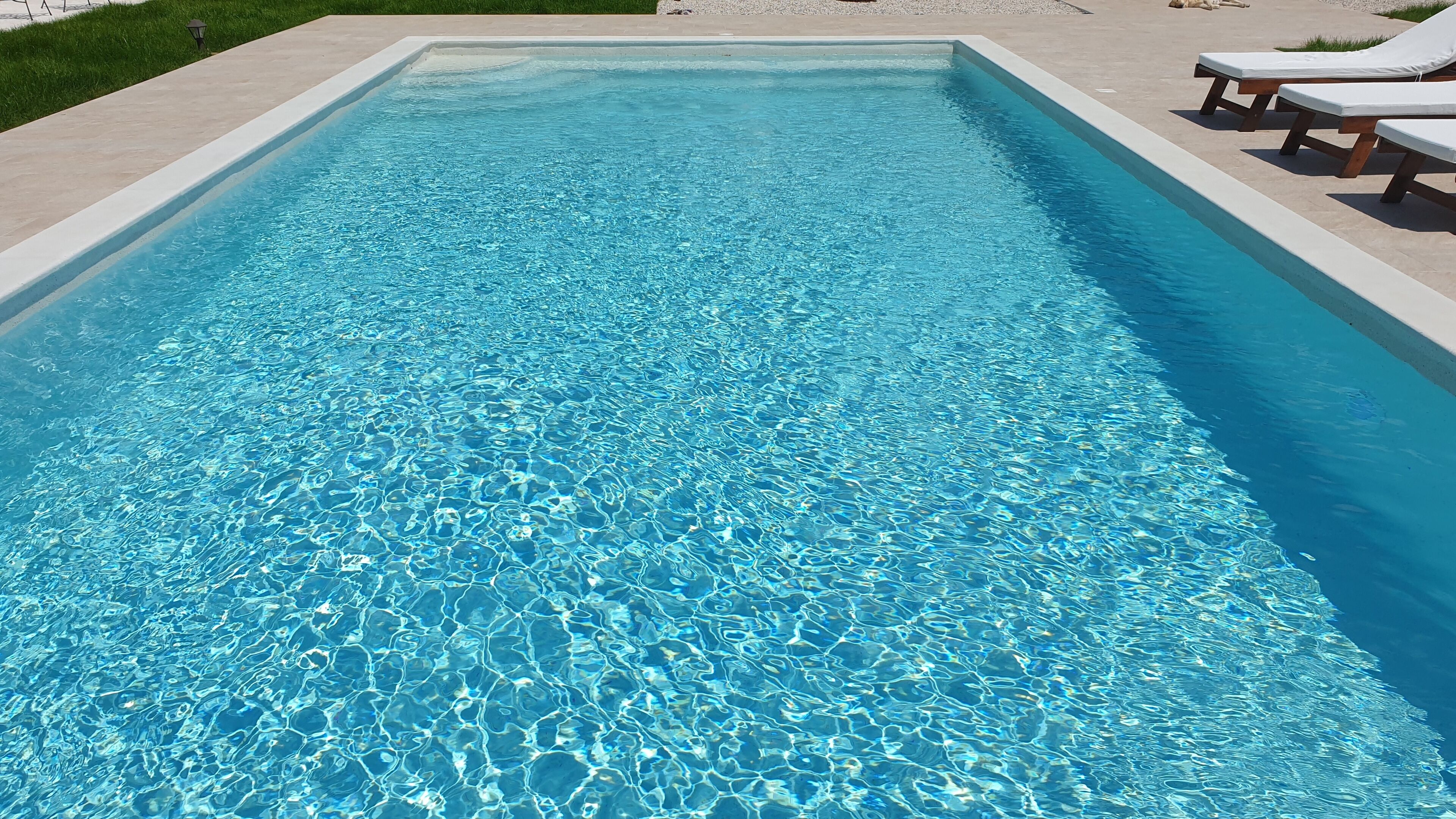 A rectangular swimming pool with clear blue water, surrounded by a light-colored deck and a few lounge chairs.