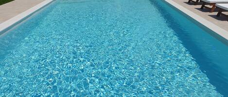 A rectangular swimming pool with clear blue water, surrounded by a light-colored deck and a few lounge chairs.