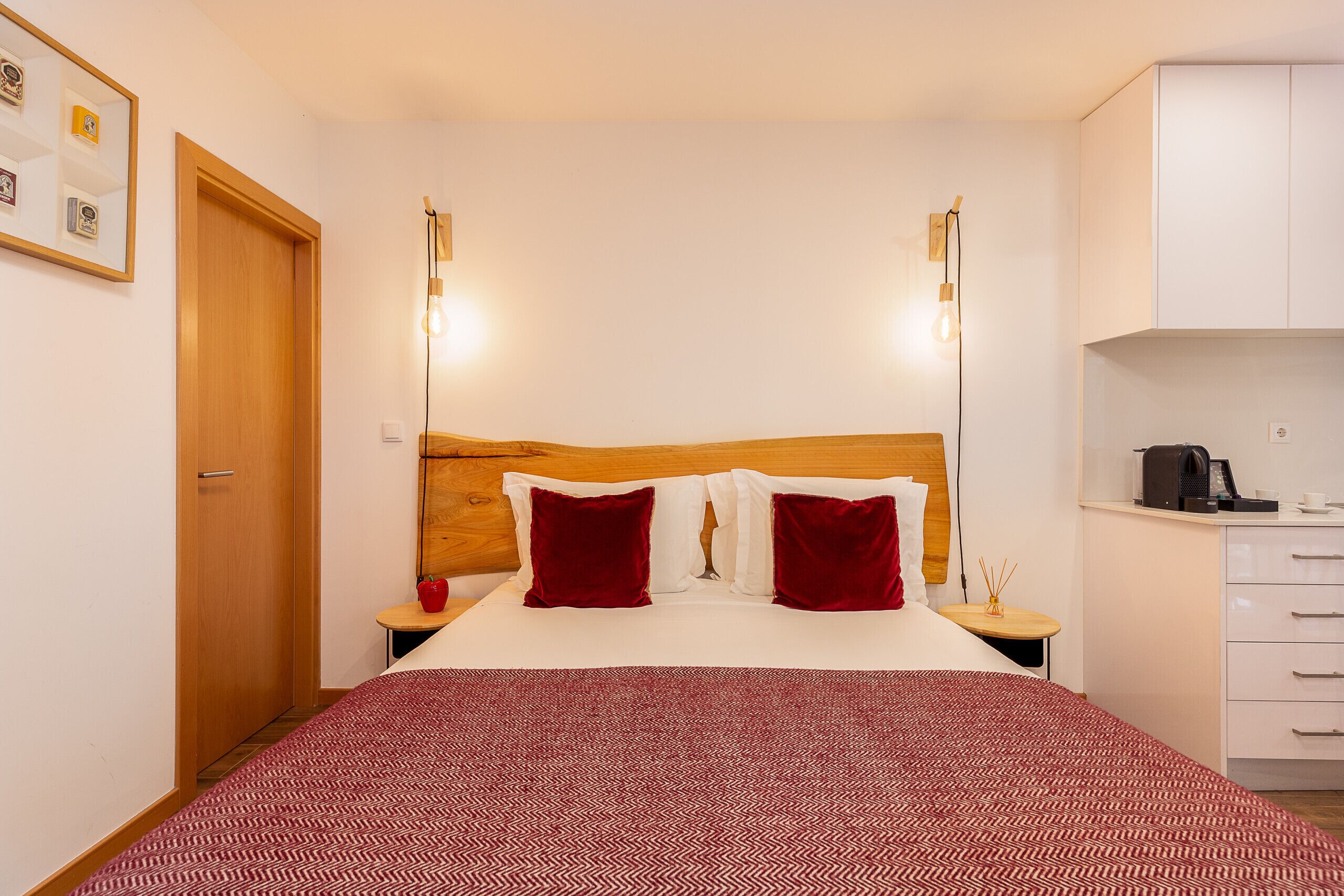 Une chambre d’hôtel bien aménagée, avec une tête de lit en bois, une couvre-lit à motifs, deux oreillers rouges, des lampes de chevet, une petite table sur laquelle se trouve un objet rouge, et un coin cuisine.