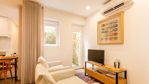 Una sala de estar moderna con un sofá, un televisor sobre un mueble de madera, una zona de comedor con una mesa y sillas, y un ventanal con cortinas.