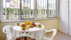 Un comedor con una mesa dispuesta para el desayuno, ventanas amplias con barrotes metálicos y vistas a un edificio y vegetación.
