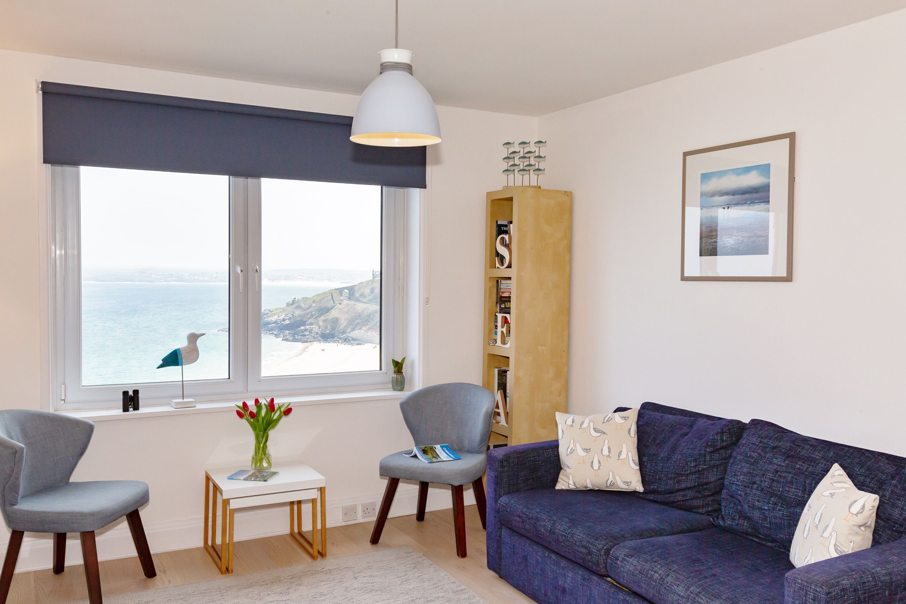 A living room with a view of the sea, a blue sofa, two chairs, a small table with a vase of flowers, and a bookshelf.