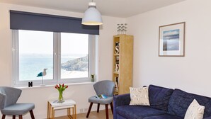 A living room with a view of the sea, a blue sofa, two chairs, a small table with a vase of flowers, and a bookshelf.