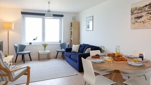 A modern living room with a blue sofa, a dining table set for breakfast, and a large window with a view of the sea.