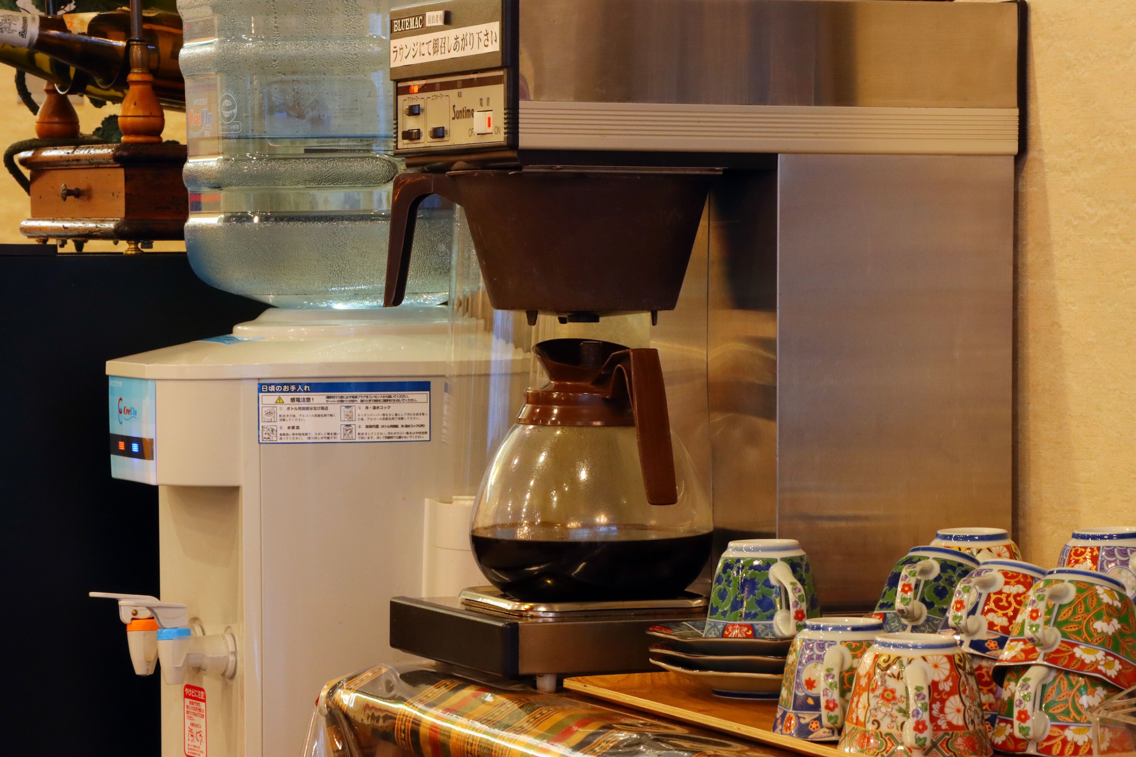 Une machine à café avec une verseuse en verre et une bouilloire, un distributeur d’eau et une collection de tasses en céramique aux couleurs vives.