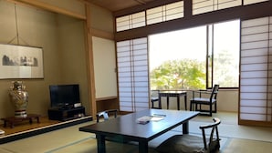 Une pièce de style japonais traditionnel, avec un sol recouvert de tatamis, une table basse entourée de chaises, un téléviseur et une grande fenêtre ornée de panneaux coulissants en papier.