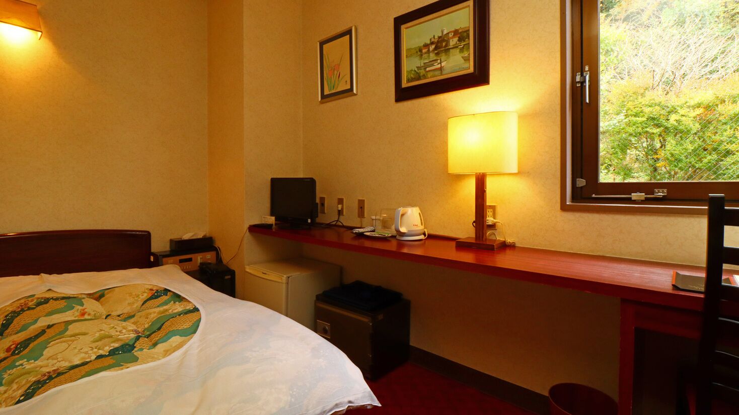 A hotel room with a bed, a desk with a lamp, a computer, and a window with a view of greenery.