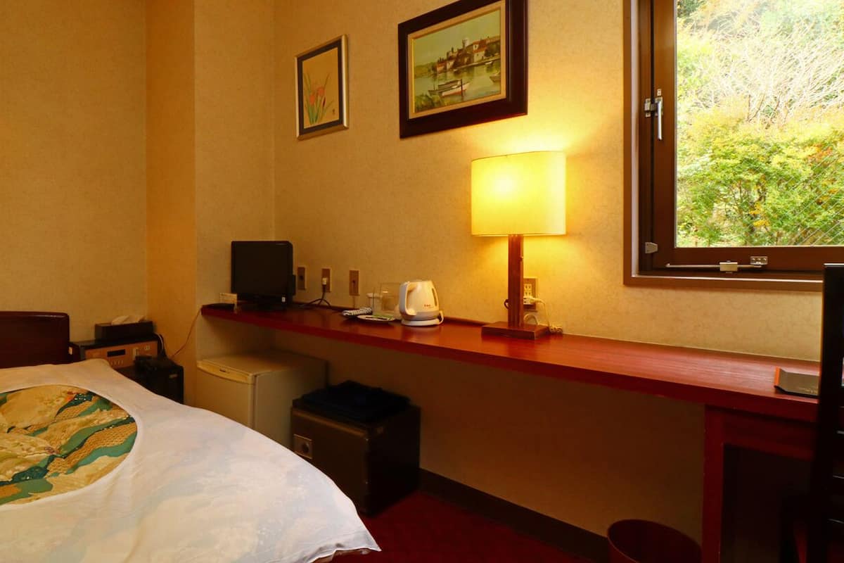 A hotel room with a bed, a desk with a lamp, a computer, and a window with a view of greenery.