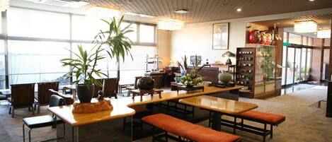 A spacious indoor area with a long wooden table and benches, surrounded by chairs and potted plants.