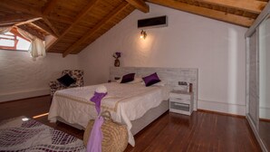 Une chambre avec un plafond en bois, un lit avec une literie blanche et des coussins violets, une table de chevet, un panier en osier et un fauteuil à motifs floraux.