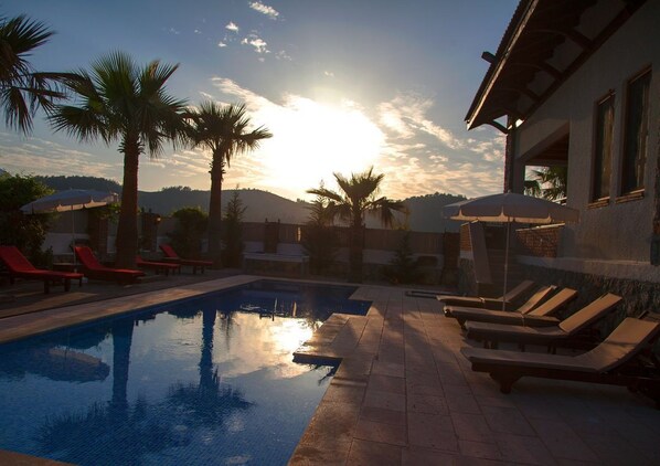 A swimming pool with lounge chairs and umbrellas, surrounded by palm trees and a building with a sloped roof. - Heated Pool - Private villa. (Fethiye)