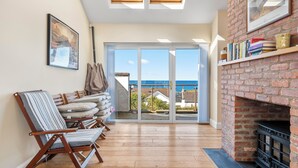 A room with a view of the sea, a fireplace, and a collection of cushions and a chair.