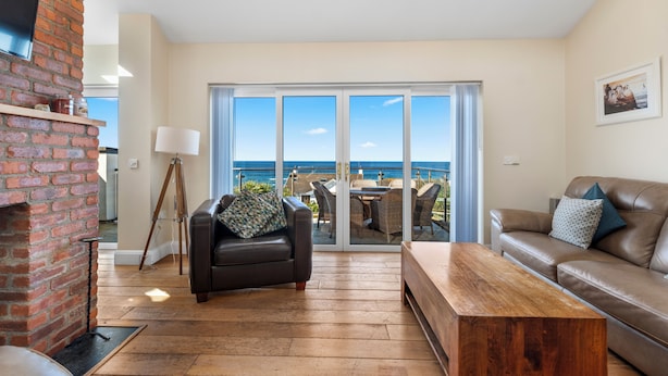 A living room with a fireplace, a sofa, a coffee table, and a balcony with a view of the ocean.