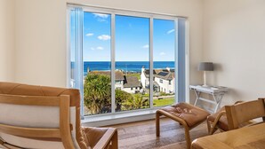 A room with a large window, a view of the sea, a wooden chair, a small table with a lamp, and a wooden bench.