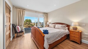 A bedroom with a wooden bed, a nightstand with a lamp, a chair, and a view of the outdoors.