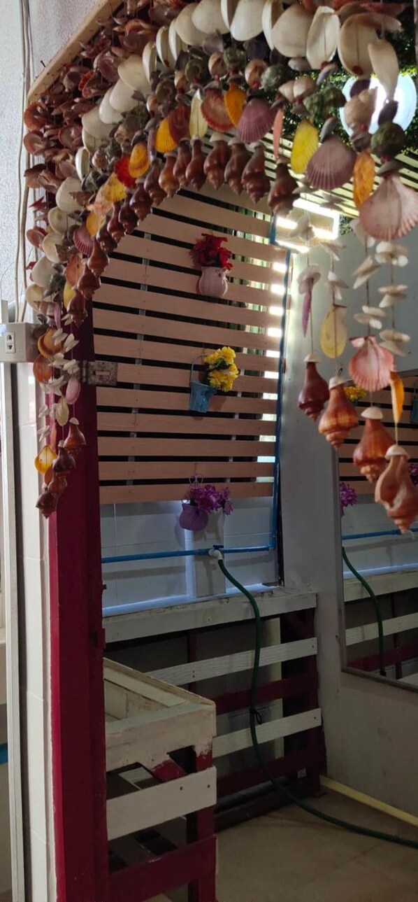 A room with a sloped ceiling decorated with hanging shells. There are two windows with blinds, and a small table with a vase of flowers. - Adchara Mansion - Hostel (Chiang Rai)
