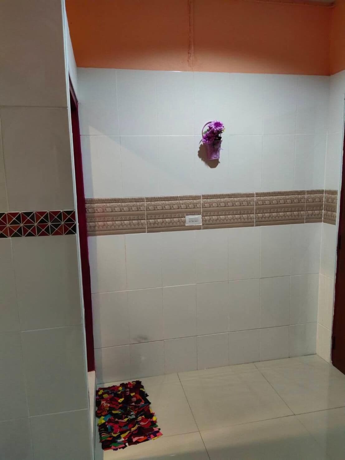 A bathroom with white tiled walls, a patterned tile border, a small purple flower decoration, and a colorful mat on the floor.