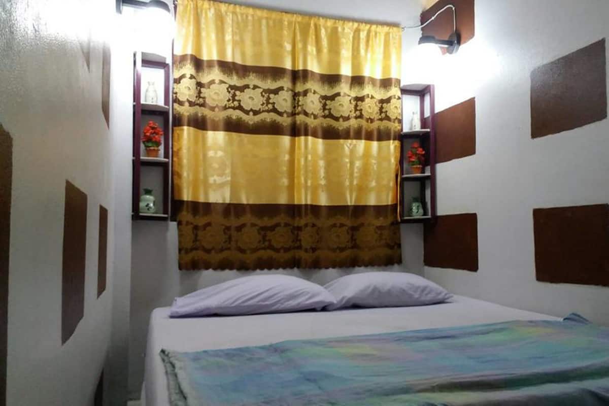 A small, single-bed room with a plain bedspread, a patterned curtain, and a wall-mounted shelf with decorative items.