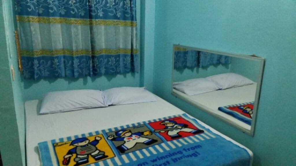 Una cama individual con colchón y almohadas blancos, manta azul y blanca a rayas con personajes de dibujos animados, una ventana con cortinas azules y un espejo en la pared.