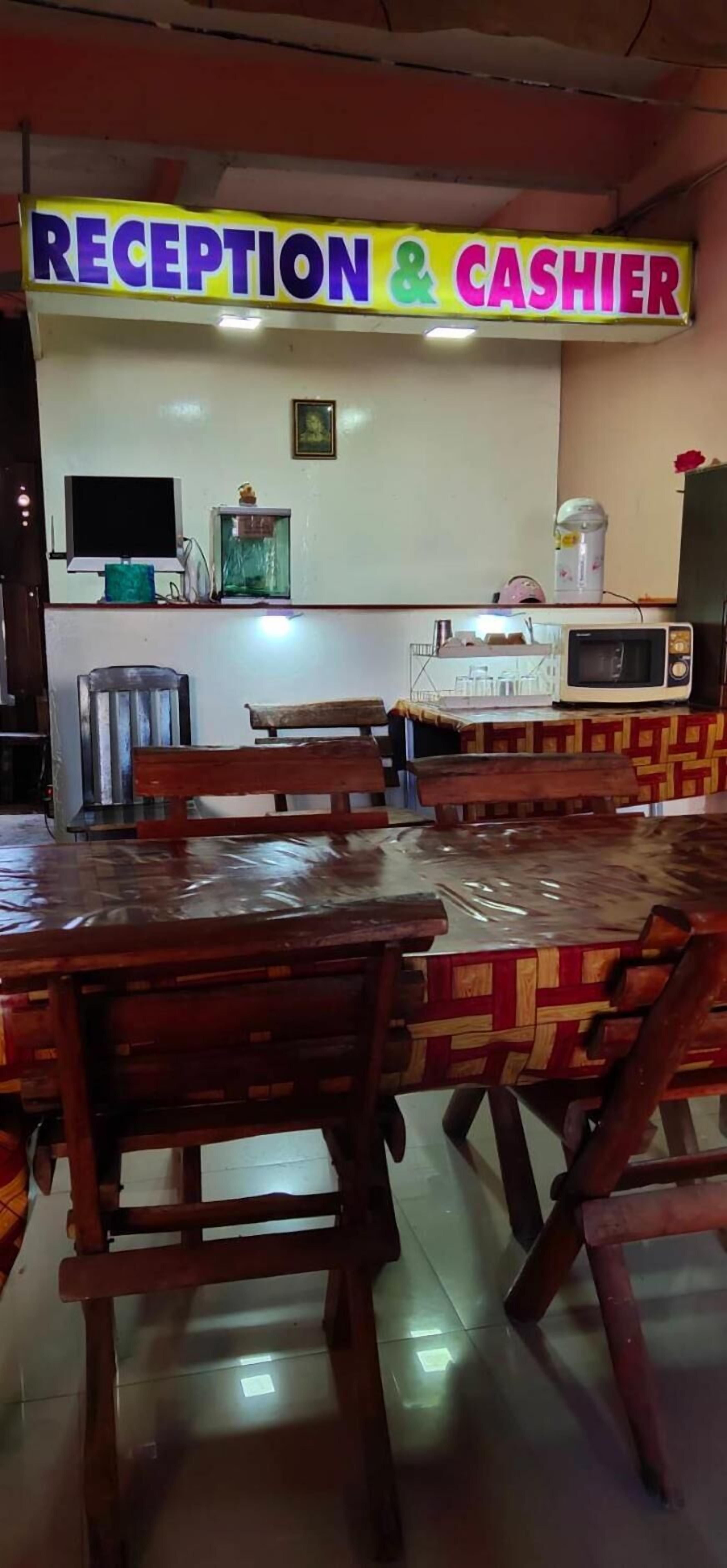 A reception area with a sign reading "RECEPTION & CASHIER" above a counter. There are wooden tables and chairs, a microwave, and a water dispenser on the counter.