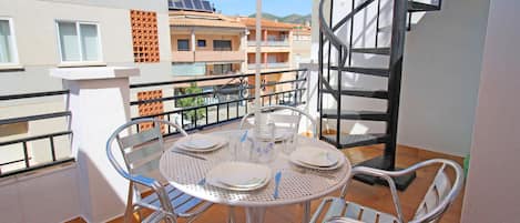 A balcony with a round metal table set for four, surrounded by metal chairs, under a striped umbrella.