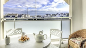 A balcony with a view of a marina and buildings, a table with a teapot, cups, and pastries, and wicker chairs.
