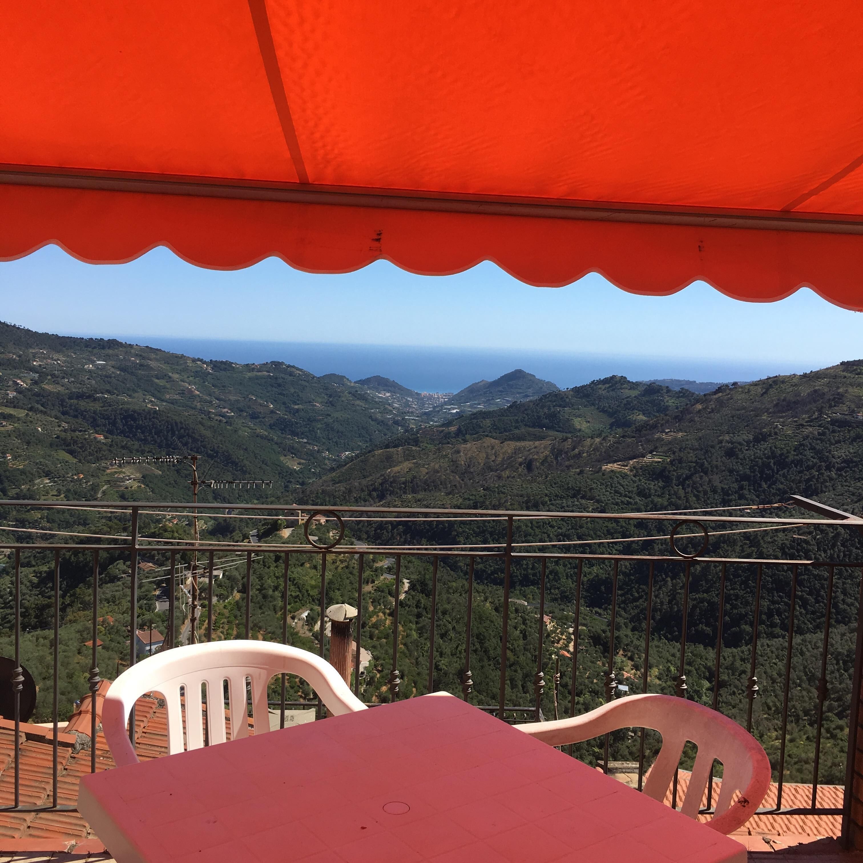 A balcony with a red canopy, white plastic chairs, and a view of a mountainous landscape with the sea in the distance.