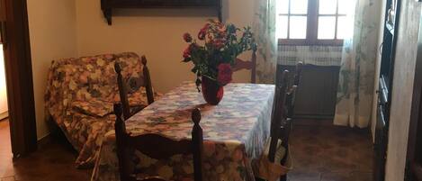 A dining area with a hexagonal tiled floor, wooden chairs, a table with a floral tablecloth, a chandelier, and a window with curtains.