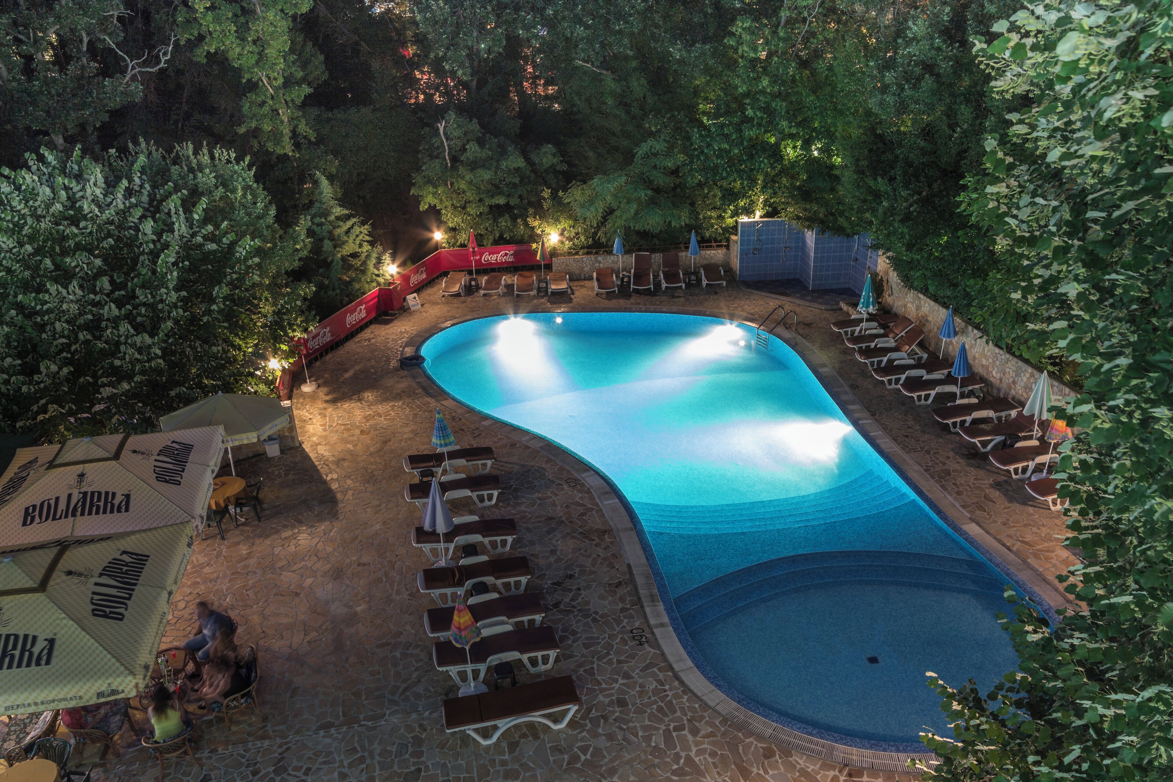 A swimming pool with lounge chairs and umbrellas, surrounded by trees and stone flooring.