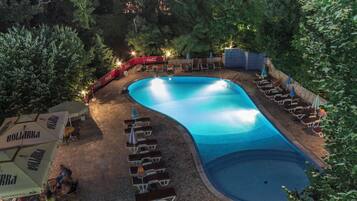 A swimming pool with lounge chairs and umbrellas, surrounded by trees and stone flooring.