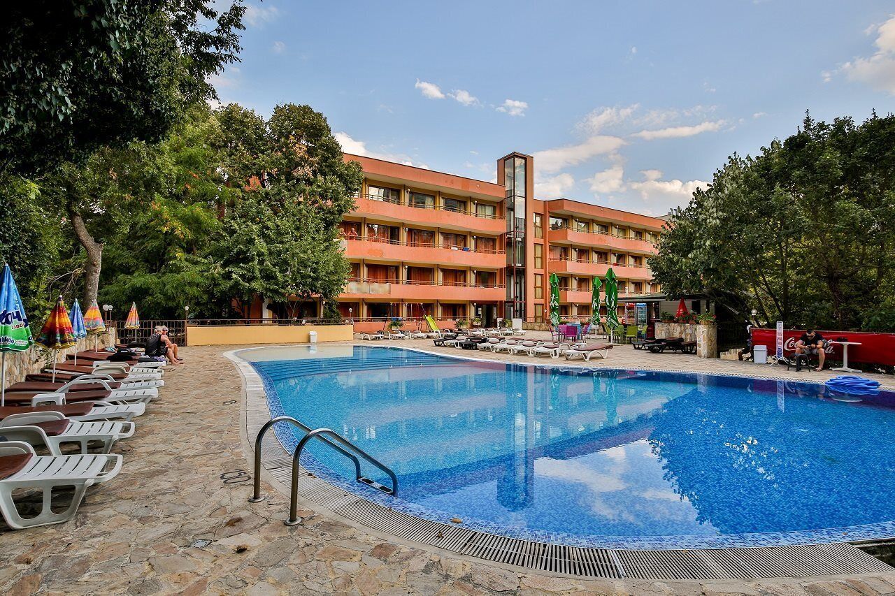 Une piscine entourée d’arbres, agrémentée de chaises longues et de parasols, et bordée d’un immeuble à plusieurs étages doté de balcons.