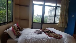 Un dormitorio con un ventanal grande, una cama con ropa de cama blanca y vistas a edificios y vegetación.