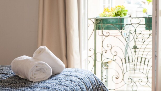 Ein ordentlich bezogenes Bett, gerollte weiße Handtücher, ein Fenster mit beigen Vorhängen und ein Balkon mit Metallgeländer und Grünpflanzen.