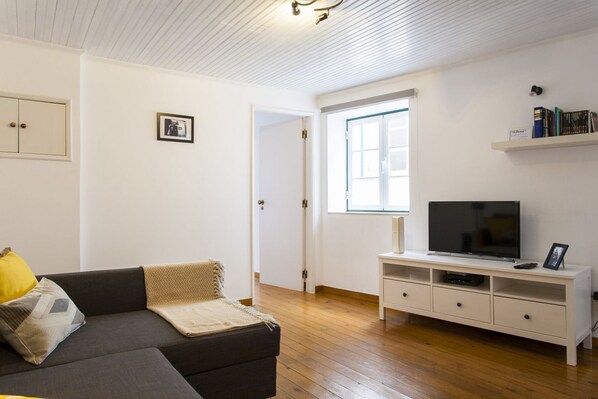Una sala de estar con un sofá, un televisor sobre un mueble blanco y una puerta que da a otra habitación.