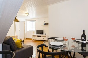 Una sala moderna con un sofá, una mesa de vidrio para comer con sillas, un televisor sobre un mueble bajo y una botella de vino sobre la mesa.