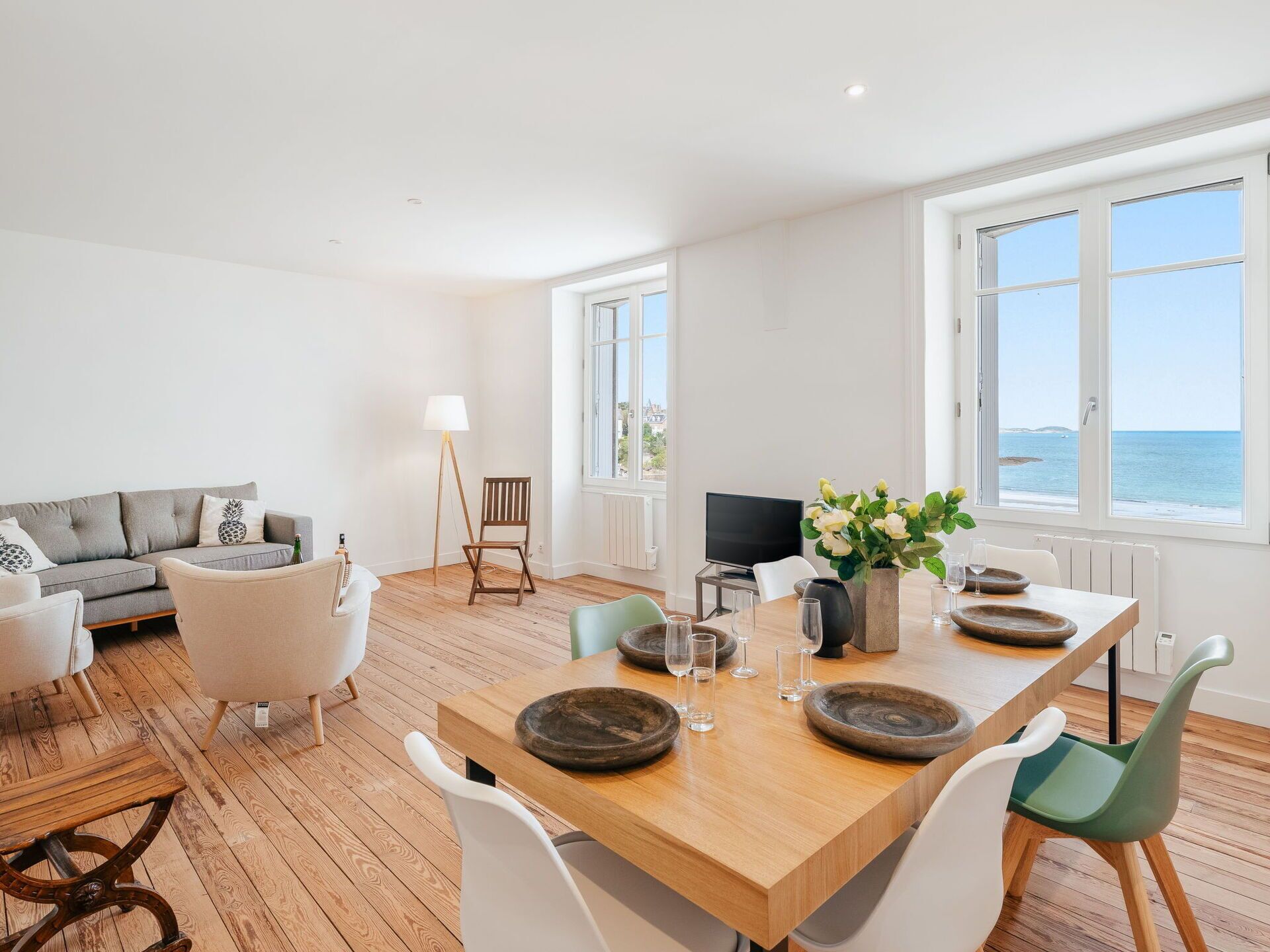 A spacious living room with a wooden dining table, white and green chairs, a grey sofa, and a TV. Large windows offer a view of the sea.