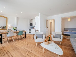 Una sala de estar moderna con piso de madera, un comedor con mesa y sillas, una cocina al fondo y un dormitorio con cama y mesita de noche.