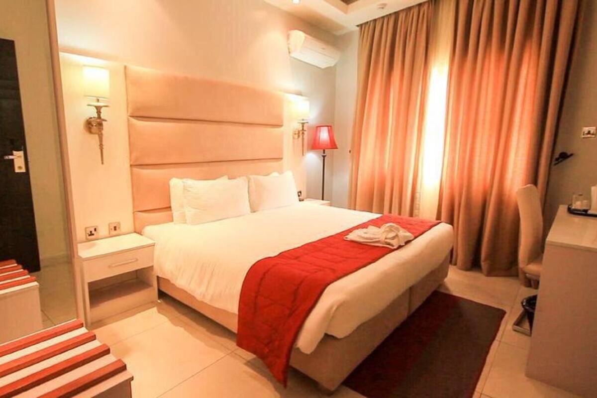 A hotel room with a large bed, a bedside table, a desk with a chair, a red throw blanket, and a window with curtains.