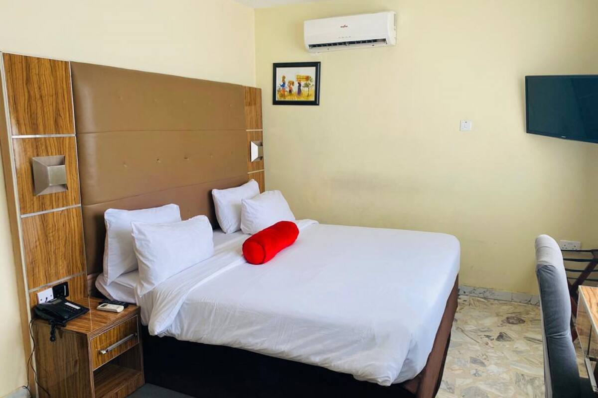 A hotel room with a large bed, a wooden headboard, a bedside table with a phone, a red pillow, an air conditioning unit, and a TV on the wall.
